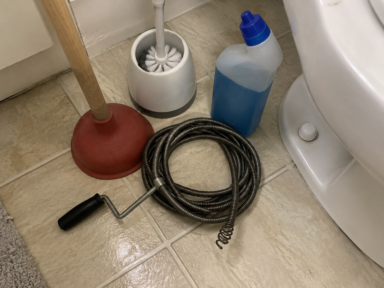 Plunger, drain snake, toilet brush and gel cleaner piled on a bathroom floor next to a toilet
